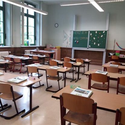 Ein klassenzimmer mit mehreren Schreibtischen und Stühlen, auf denen Bücher und Materialien liegen, eine grüne Tafel mit 'Herzlich Willkommen' und hellem Tageslicht, das durch große Fenster einfällt.